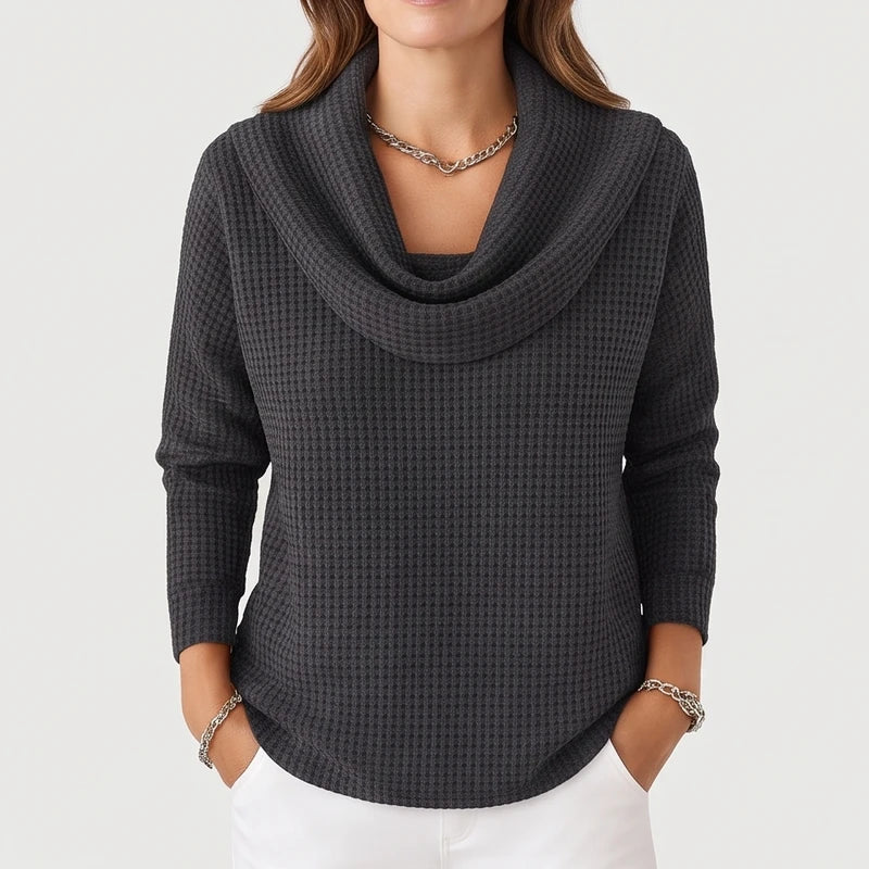 Woman wearing a charcoal gray waffle knit cowl neck sweater with a silver chain necklace and white pants, hands in pockets.
