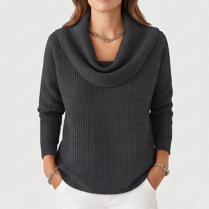 Woman wearing a charcoal gray waffle knit cowl neck sweater with a silver chain necklace and white pants, hands in pockets.