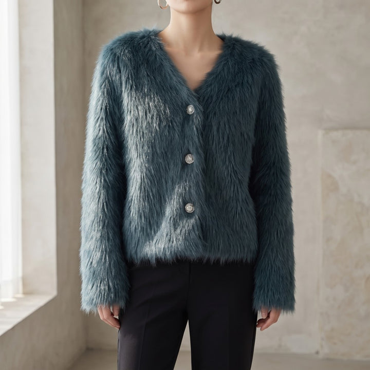 A woman wearing a teal blue faux fur cardigan with silver buttons, paired with black pants, standing in a softly lit interior setting.
