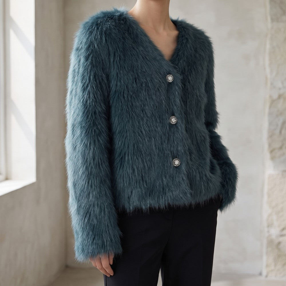 A person wearing a fuzzy long-sleeve cardigan in a muted blue-gray color with three silver buttons, paired with black pants, standing in a softly lit indoor setting.