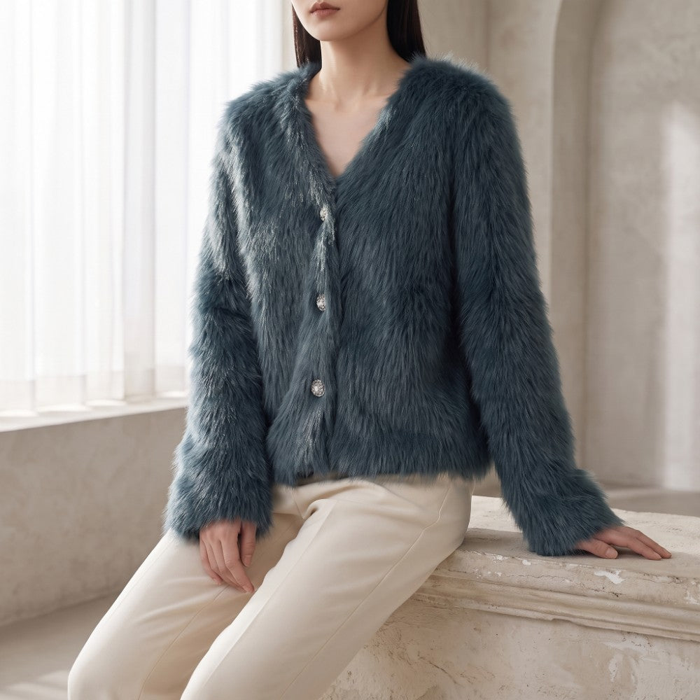 A woman wearing a plush, textured blue-gray faux fur cardigan with silver buttons, paired with cream-colored pants, sitting on a stone ledge in a softly lit interior setting.