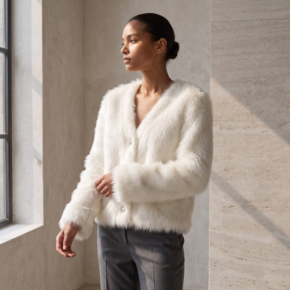A woman wearing a fluffy white faux fur cardigan with a V-neck and button closure, paired with dark gray trousers, standing in a modern room with textured beige walls and a window.