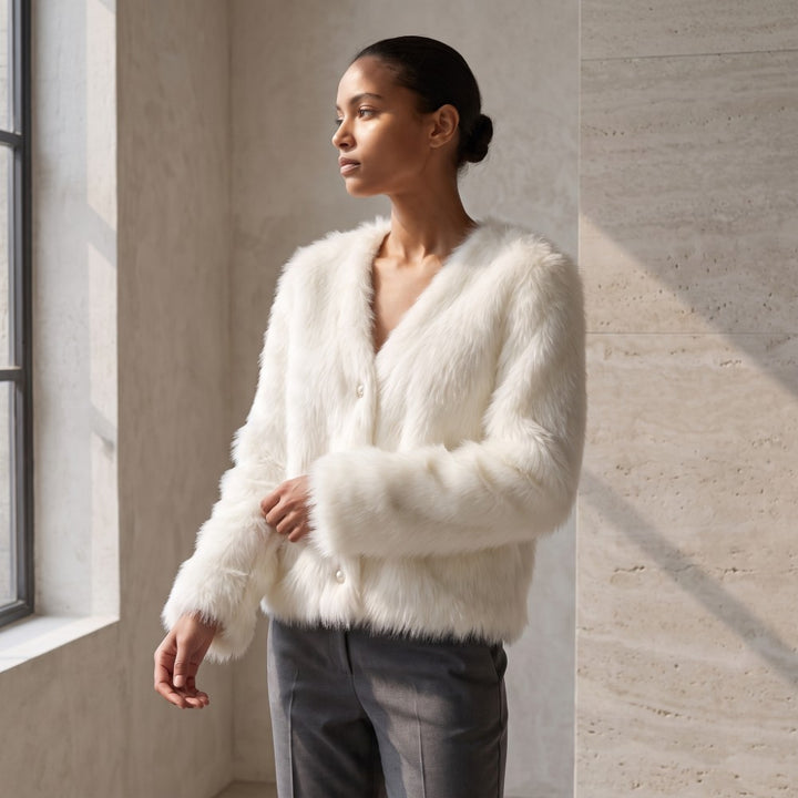 A woman wearing a fluffy white faux fur cardigan with a V-neck and button closure, paired with dark gray trousers, standing in a modern room with textured beige walls and a window.