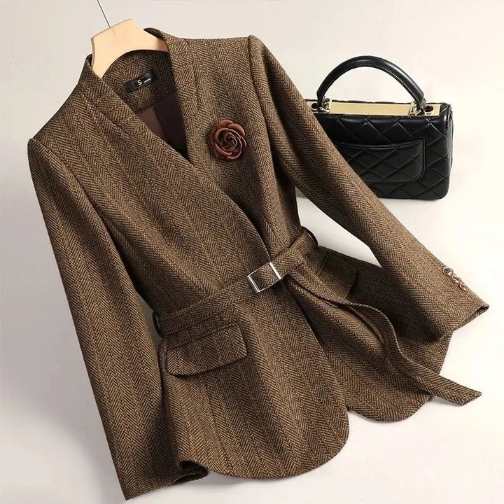 Brown herringbone blazer with a belted waist, front flap pocket, flower brooch, and visible cuff buttons, beside a black quilted handbag.