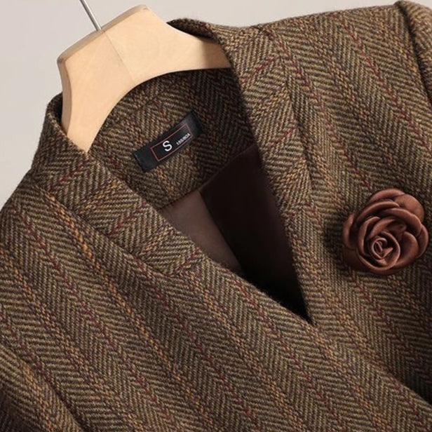 Close-up of a brown herringbone blazer with a satin rosette lapel pin, hanging on a wooden hanger.