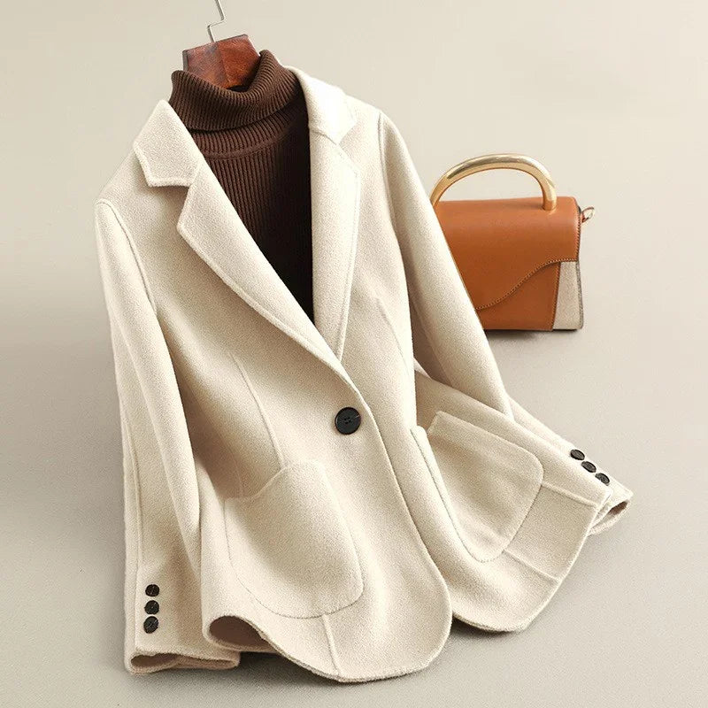 Cream colored short coat with a single black button closure, large front pockets, and a ribbed brown turtleneck visible underneath.