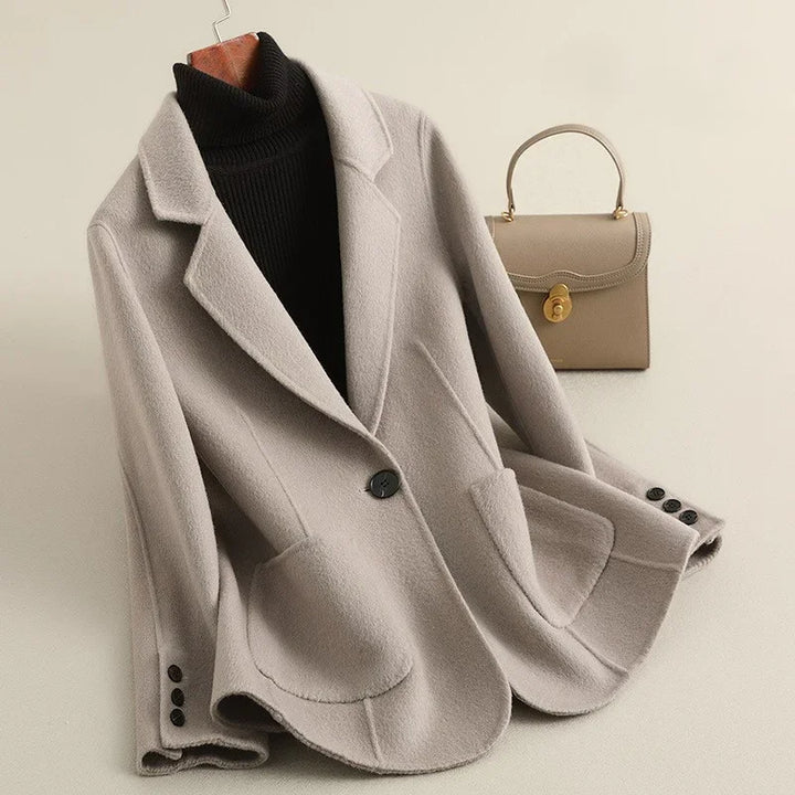 Light gray wool blazer on a hanger with a black turtleneck, featuring a single black button and two rounded pockets.