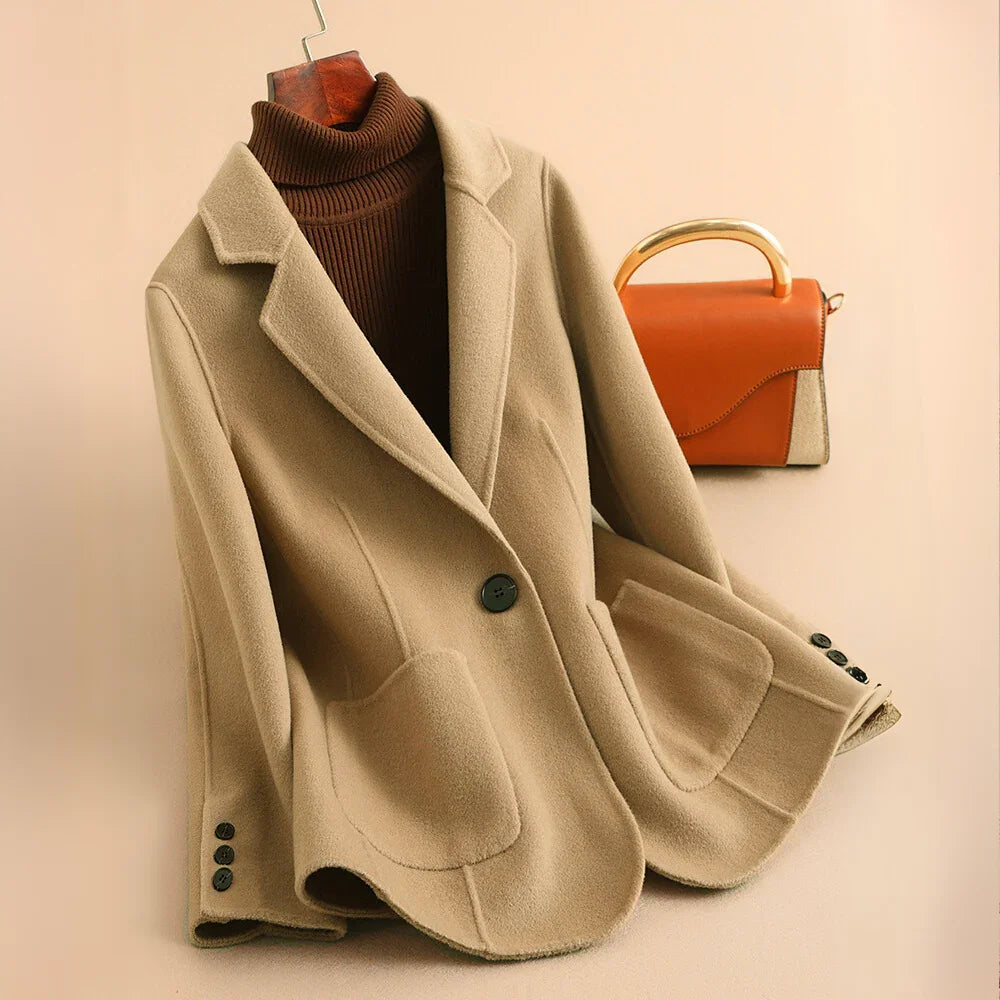 Light beige tailored short coat with large lapels, one black button closure, and two rounded patch pockets, with a brown turtleneck and a brown and gold handbag in the background.