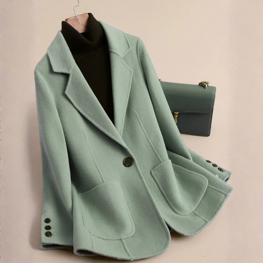 Light sage green blazer-style wool coat with large lapels, a single button, and two curved patch pockets, hung on a hanger.
