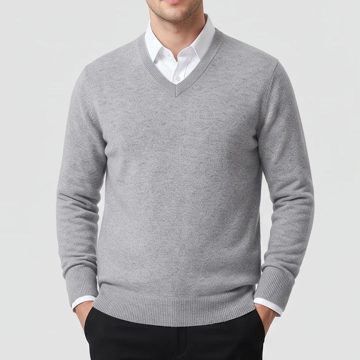 Man wearing a light grey v-neck knit sweater layered over a crisp white collared shirt, posing with hands in pockets.