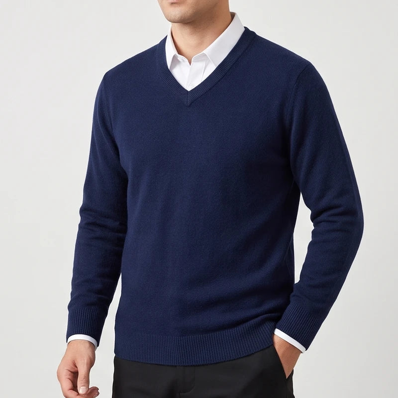 Man wearing a navy blue V-neck sweater layered over a white collared shirt with black trousers against a plain background.