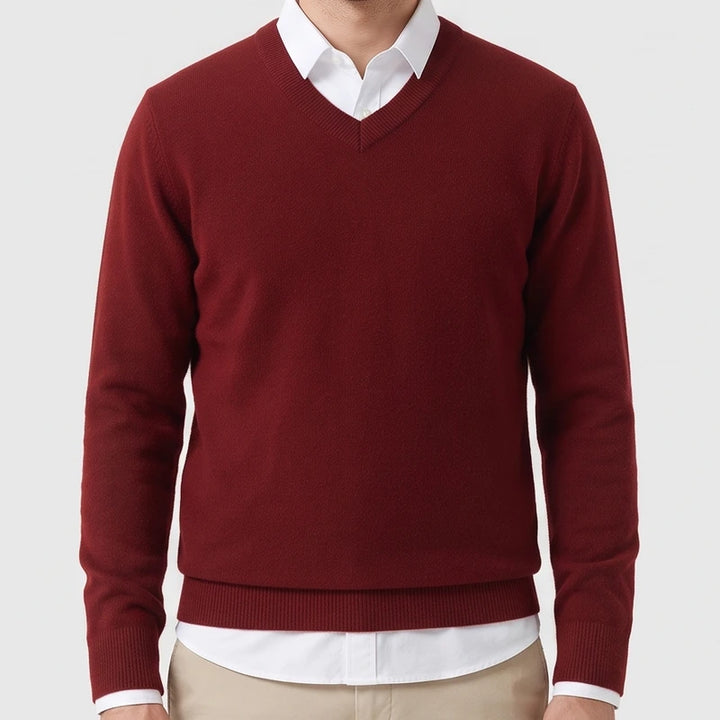 Man wearing a burgundy knit V-neck sweater layered over a white collared shirt and beige trousers against a light background.