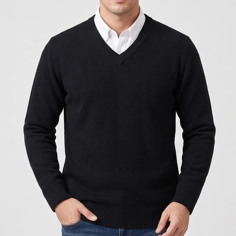 Man wearing a black V-neck knit sweater layered over a white collared shirt with blue jeans against a light background.