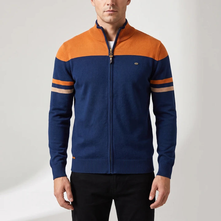 Man wearing a navy and orange zip-up sweater with two horizontal beige stripes on the left sleeve, standing on a white background.