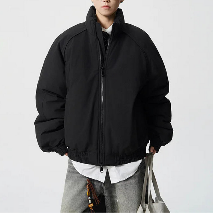 A man wearing a black puffer jacket with a high collar and front zipper, styled over a white shirt and gray pants, standing against a light gray studio background.