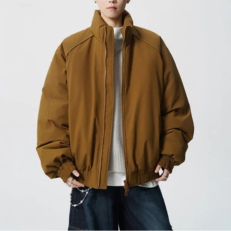 A young man modeling an oversized brown puffer jacket with a high collar, zippered front, and elastic cuffs, styled over a white sweater and dark blue jeans against a light gray studio background.