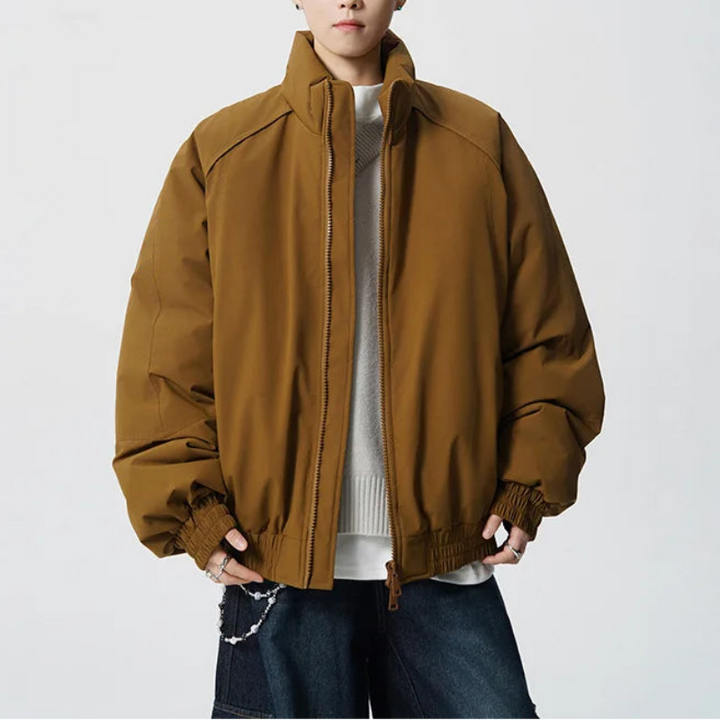 A young man modeling an oversized brown puffer jacket with a high collar, zippered front, and elastic cuffs, styled over a white sweater and dark blue jeans against a light gray studio background.