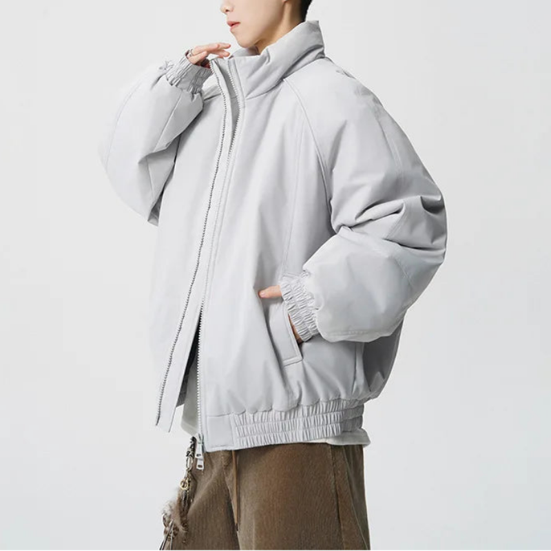 A model wearing a light gray oversized zip-up jacket with a high collar and elastic cuffs, paired with brown pants, set against a plain light gray background.