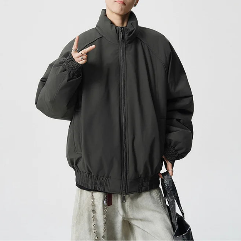 A person modeling a dark charcoal gray zip-up jacket with a high collar, elastic cuffs, and a relaxed fit, paired with light gray pants and black sunglasses against a plain light gray background.