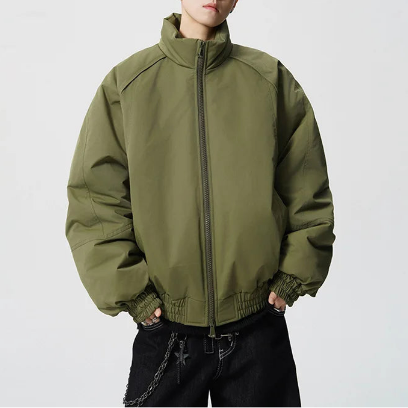 A model wearing an oversized olive green zip-up jacket with a high collar, paired with black pants and sunglasses, set against a plain light gray background.