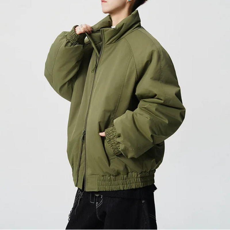 A model wearing an oversized olive green puffer jacket with elasticized cuffs and hem, styled with black pants against a light gray studio background.