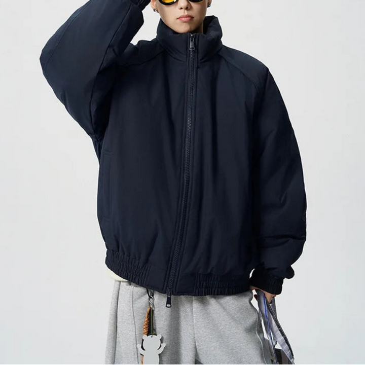 A man wearing a dark navy blue zip-up puffer jacket, styled with gray sweatpants and yellow-tinted sunglasses, standing against a light gray studio background.