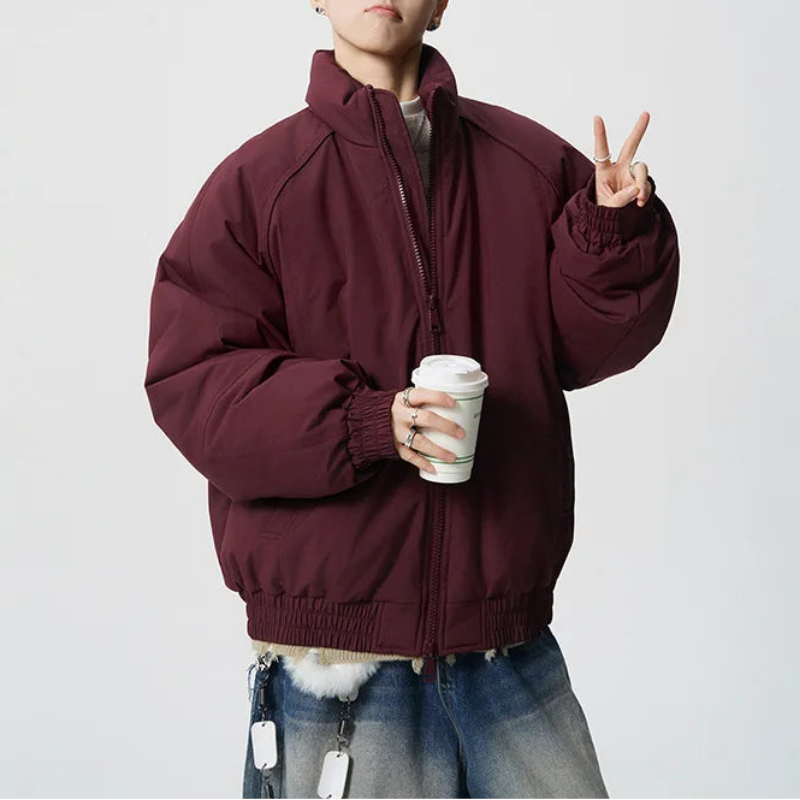 A person wearing an oversized maroon zip-up puffer jacket, styled with blue jeans and holding a white coffee cup, posing against a light gray studio background.