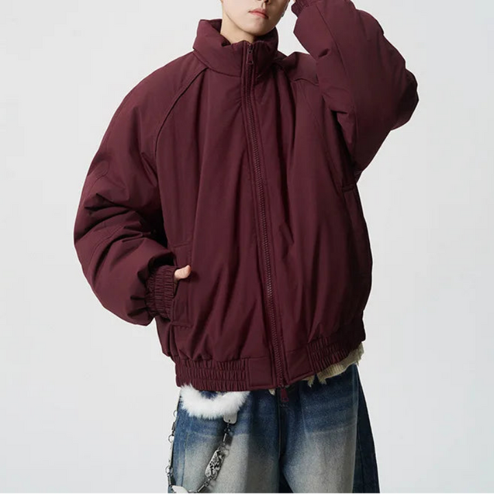 A person modeling an oversized maroon zip-up jacket with a high collar, paired with blue jeans and sunglasses, against a plain light gray background.